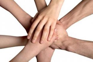 #PraCegoVer: Foto mostra sete mãos unidas em um feixe de braços, vistas de cima, um símbolo de união. 