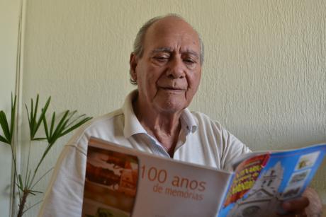 #PraCegoVer: Foto mostra o ex-vereador Laércio Conte lendo uma revista comemorativa. Na capa da revista, está a foto da Matriz de São Sebastião e atrás está escrito 100 anos de memórias.