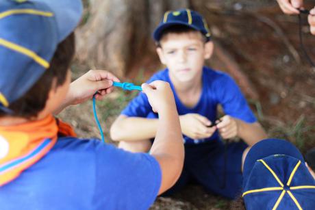 #PraCegoVer - Um menino vestido com roupas de escoteiro mostra ao outro como fazer um nó com um cordão azul