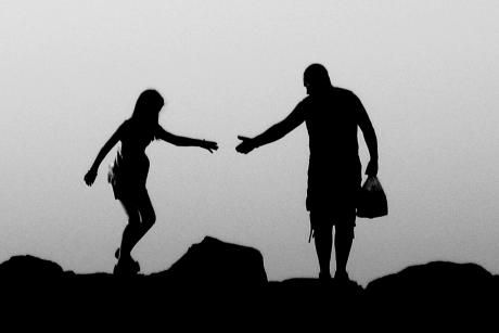 #PraCegoVer: Foto mostra silhueta de rapaz e moça caminhando no topo de uma montanha ou morro. O homem segura um saco e estende a mão para ajudar a mulher a atravessar um desnível do terreno.